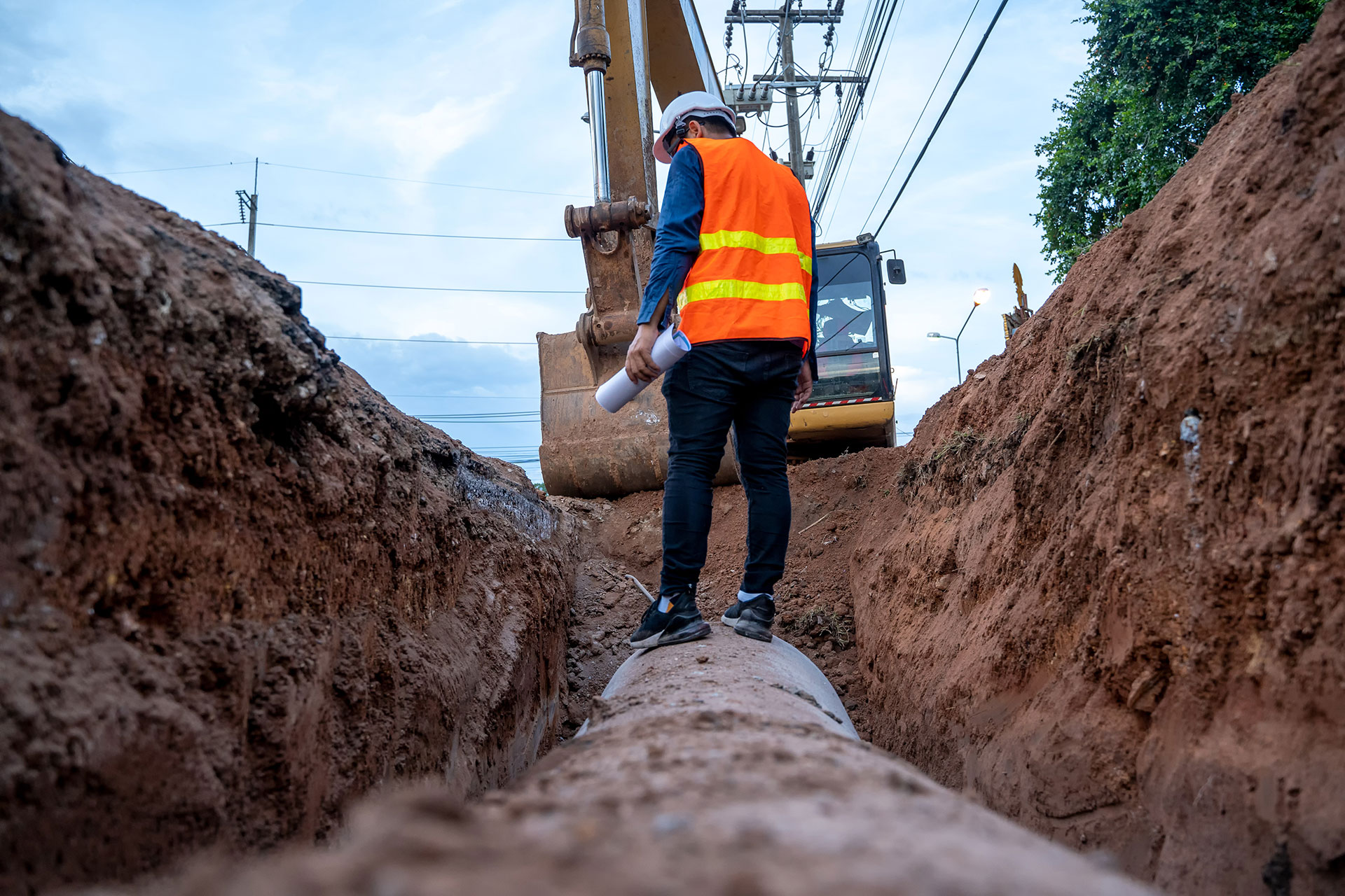 A Southern Water engineer stands on top of a pipe that has been excavated from underground. There is a digger behind them 